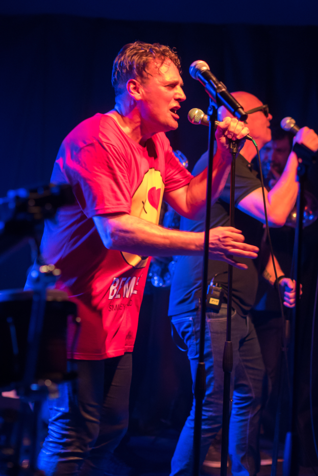 Ein Mann in einem roten Hemd und Brille singt auf der Bühne in ein Mikrofon, umgeben von Musikern, die Instrumente spielen.