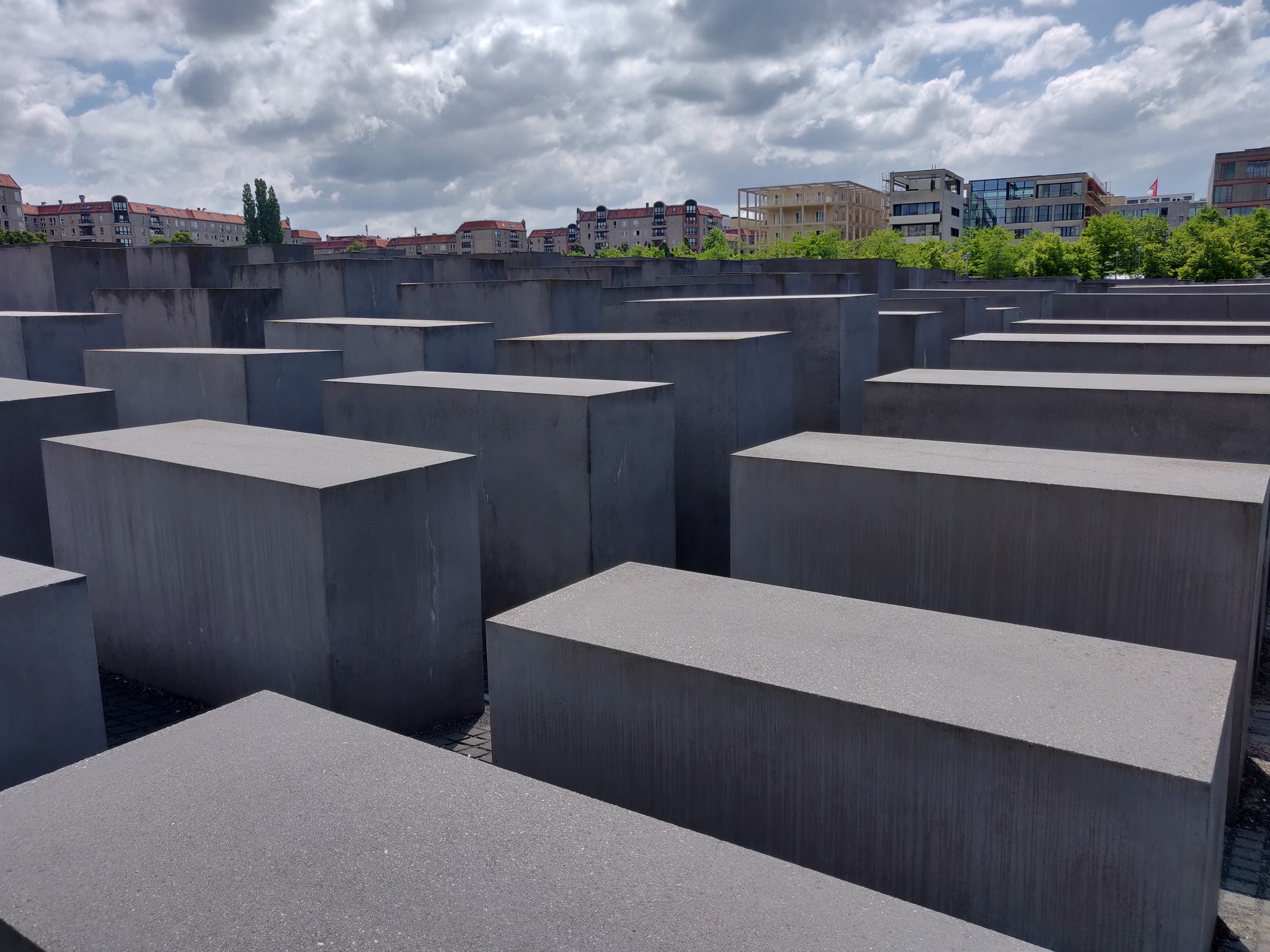 Betonschwellen in einem Gittermuster am Holocaust-Mahnmal für die ermordeten Juden Europas in Berlin, mit Bäumen, Gebäuden und Wolken im Hintergrund.