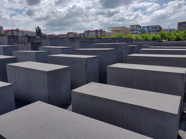 Betonschwellen in einem Gittermuster am Holocaust-Mahnmal für die ermordeten Juden Europas in Berlin, mit Bäumen, Gebäuden und Wolken im Hintergrund.