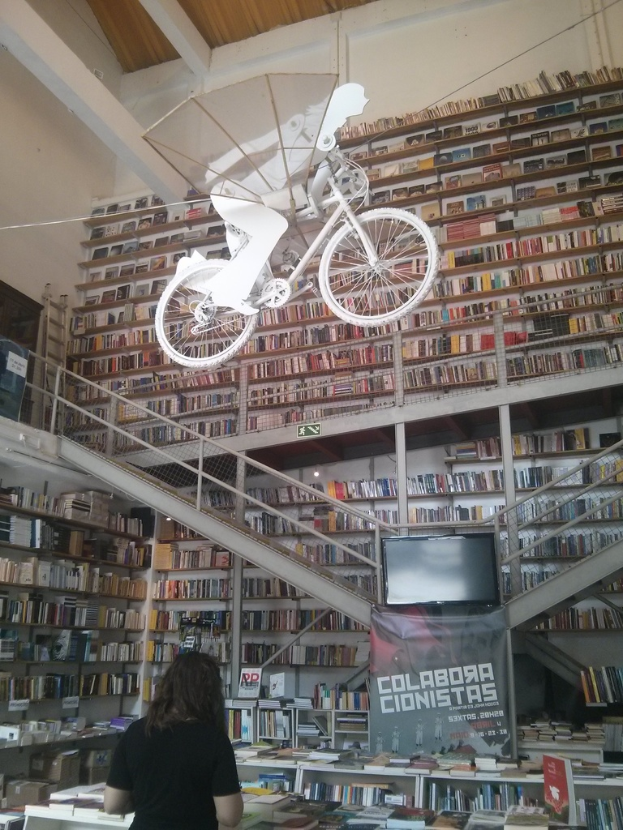 Ein Innenblick in eine Bibliothek mit auf Regalen und Tischen angeordneten Büchern und einer Person, die mit dem Rücken zur Kamera steht, einem Bildschirm mit einem Banner, einem Geländer und einer Treppe sowie einem Schild, das eine Person auf einem Fahrrad zeigt.