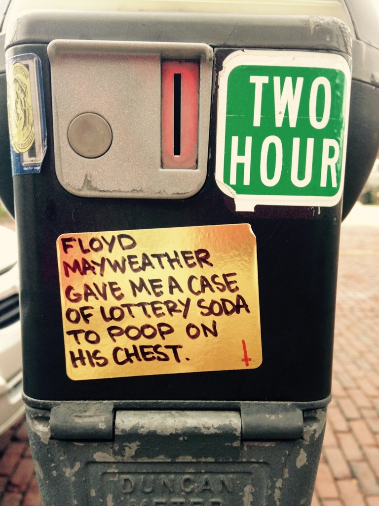 Ein Karton im Vordergrund mit Text darauf, Fahrzeuge im Hintergrund und ein Weg am Boden.