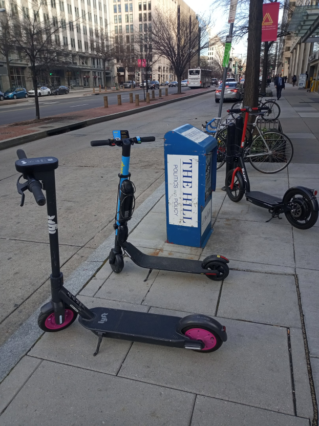 Zwei Elektro-Scooter auf einer städtischen Straße geparkt mit Fahrrädern, einem Müllcontainer, Fahrzeugen, Bäumen, Gebäuden und Menschen im Hintergrund.