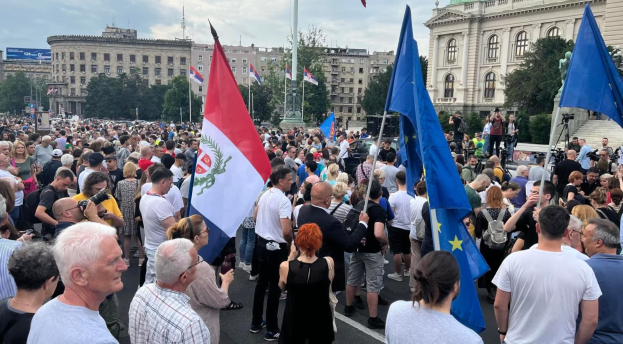 Eine große Gruppe von Menschen steht vor einem Gebäude, hält Fahnen und Kameras, mit Bäumen und Laternen im Hintergrund unter einem bewölkten Himmel, wahrscheinlich protestierend in Budapest, Ungarn.
