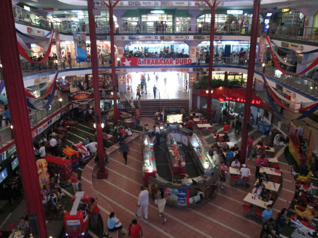 Ein belebter Einkaufszentrum-Szenen mit Säulen, Menschen, Tischen, Fliesen, Flaggen, Geschäften, einem Banner, Treppen und einem Namensschild.