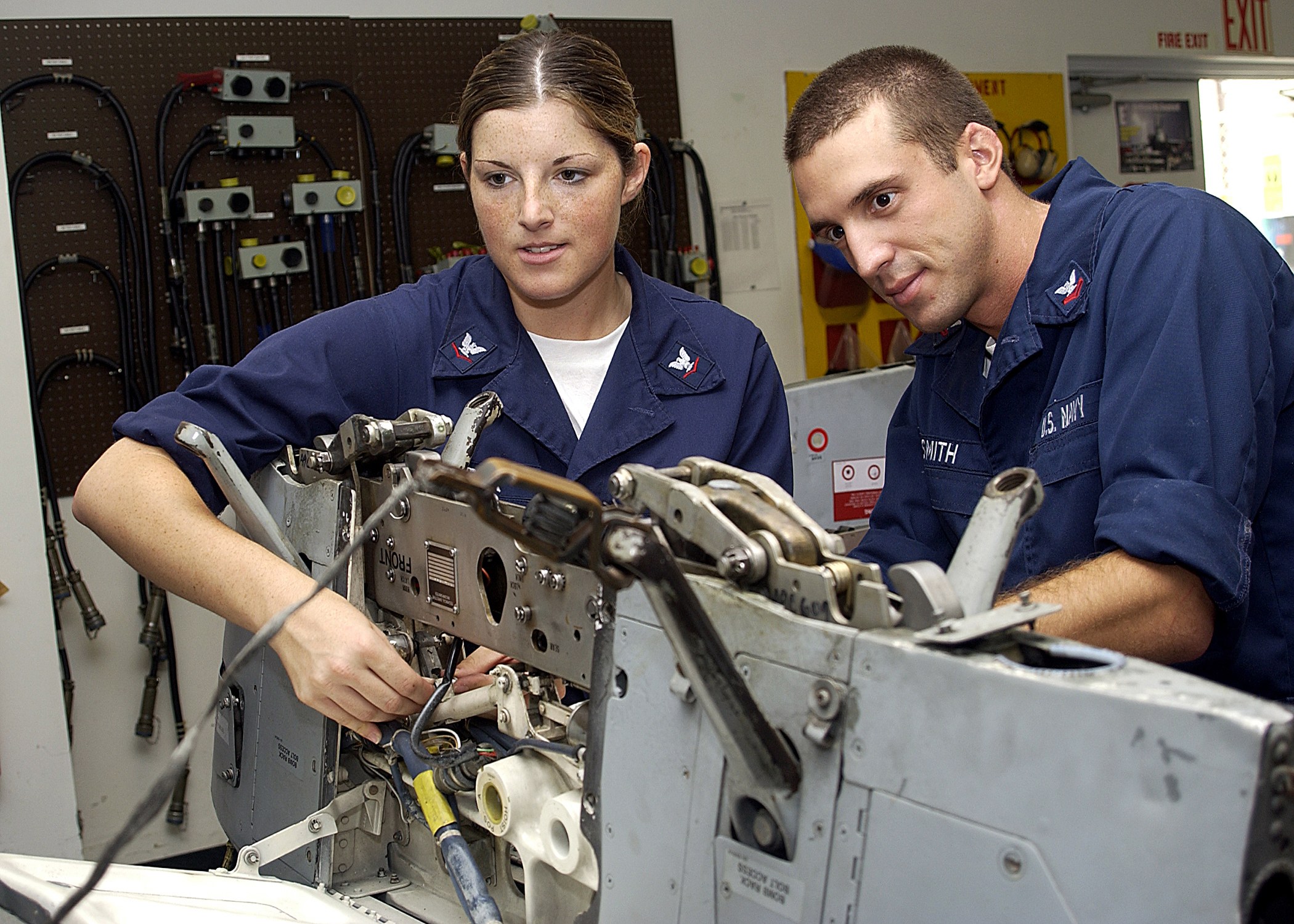 Zwei Fabrikarbeiter in blauen Uniformen bedienen Maschinen, mit industrieller Ausstattung und einer Wandtafel im Hintergrund.