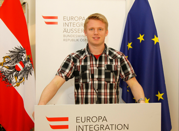 Ein Junge steht hinter einem Podium mit einem Mikrofon, mit einem Plakat und Fahnen dahinter.