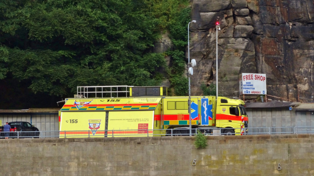 Ambulanz am Straßenrand neben einer Klippe geparkt, mit verschiedenen Objekten im Vordergrund und Bäumen und einem Auto im Hintergrund.