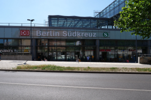 Großes gläsernes Berliner Südkreuz-Bahnhof mit sichtbarem Text, umgeben von Straßenlaternen, Lichtern, Fahrzeugen, Fußgängern, Fahrrädern, Bäumen und einem klaren blauen Himmel.