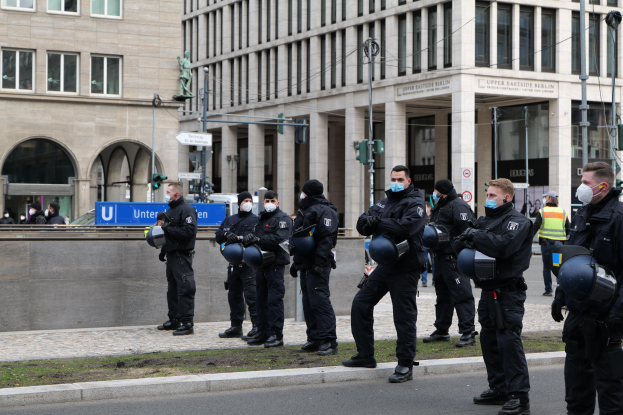 Eine Gruppe von Polizisten in schwarzen Uniformen und Masken steht vor einem Gebäude mit Glasfenstern und Säulen in Berlin, Deutschland, einige halten Helme, mit Laternenmasten, Verkehrsampeln, Schildern mit Text, einer Statue, anderen Menschen und grasbewachsenem Boden im Hintergrund.