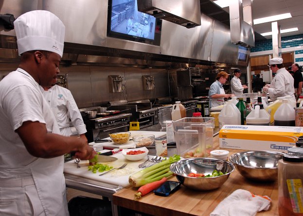 Eine Gruppe von Köchen in einer Küche, mit einem im Vordergrund in einer weißen Schürze und Mütze, der ein Messer hält und von verschiedenen Utensilien, Schüsseln, Gemüse, einem Handy, Flaschen und anderen Gegenständen umgeben ist, während im Hintergrund ein Fernseher zu sehen ist.