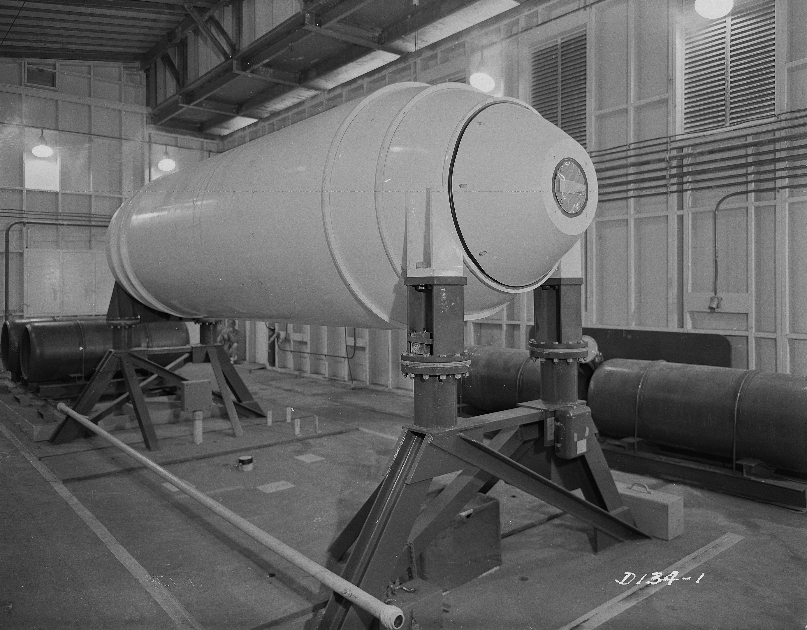 Große zylindrische Wasserstoffbombe auf einem Ständer in einer industriellen Fabrikumgebung mit sichtbaren Rohren, Lampen und Text am unteren Bildrand.