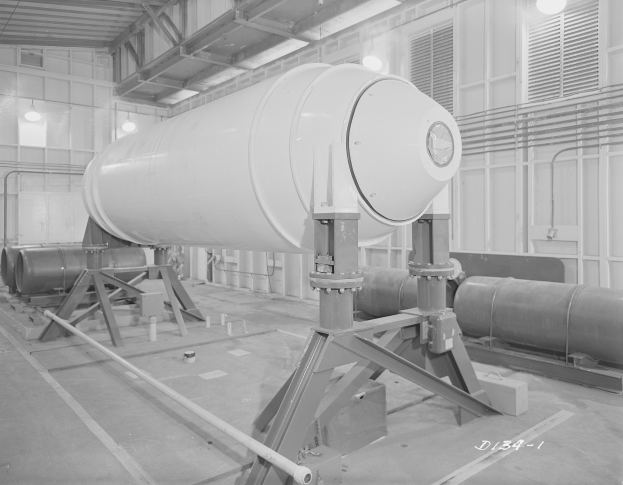 Große zylindrische Wasserstoffbombe auf einem Ständer in einer industriellen Fabrikumgebung mit sichtbaren Rohren, Lampen und Text am unteren Bildrand.