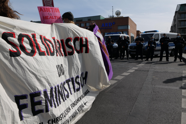 Gruppe von Menschen marschiert auf einer Straße mit einem Banner 'Solidarität und Feminismus' vorbei an parkenden Autos und Gebäuden unter einem klaren blauen Himmel.