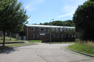 Ein Schulgebäude mit Fenstern, einem Straßenschild, einem Schild und einer Gruppe von Bäumen, umgeben von Gras und einem Zaun, unter einem bewölkten Himmel.