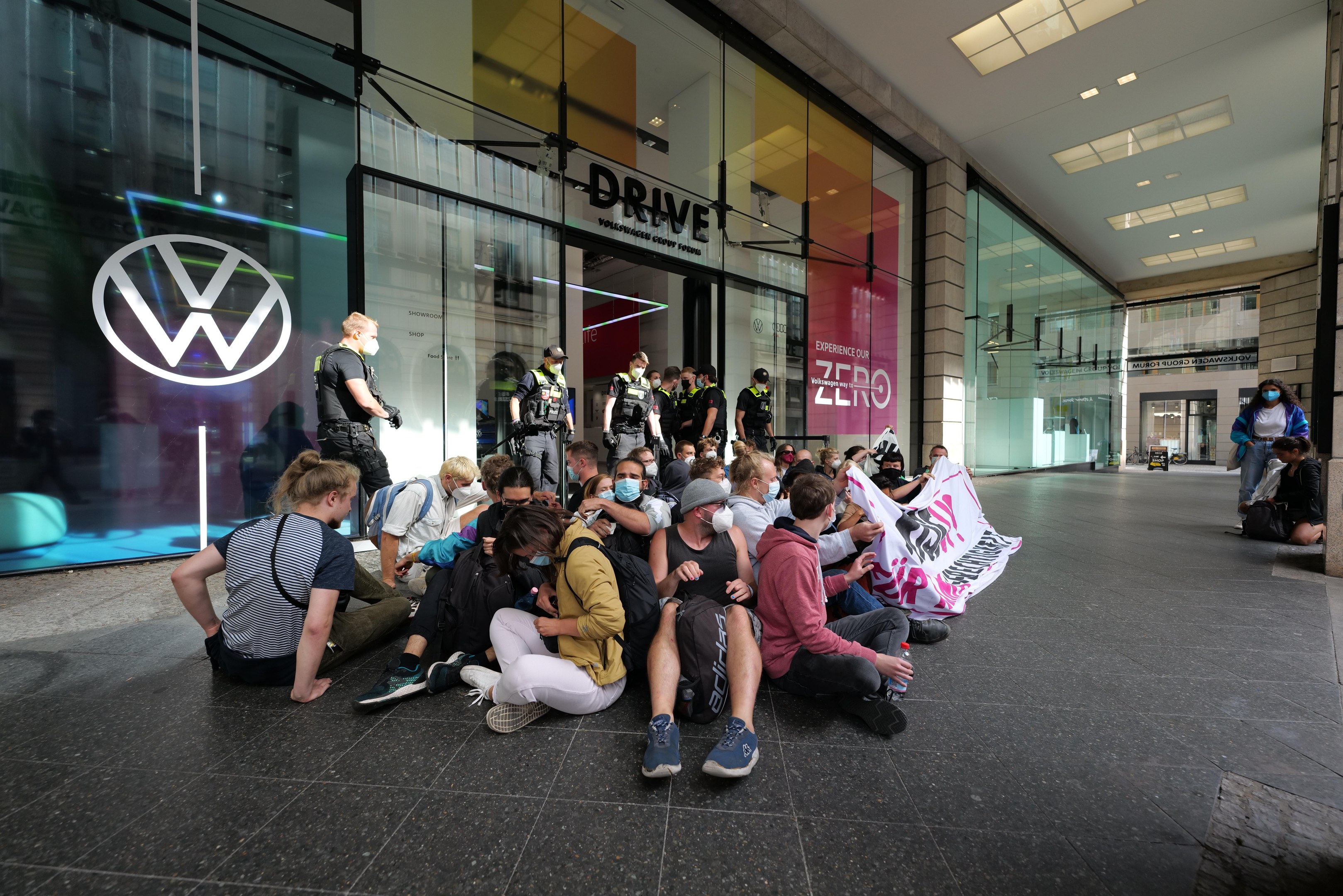 Eine Gruppe von Menschen sitzt vor einer Volkswagen-Vertretung mit Glaswänden auf dem Boden und hält eine Fahne mit Text, unter Deckenlampen, scheinbar protestierend.