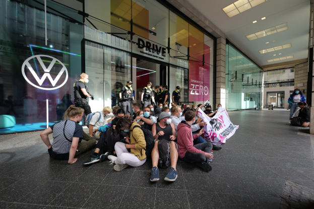 Eine Gruppe von Menschen sitzt vor einer Volkswagen-Vertretung mit Glaswänden auf dem Boden und hält eine Fahne mit Text, unter Deckenlampen, scheinbar protestierend.