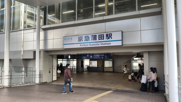 Menschen mit Gepäck durch eine Bahnhofshalle gehen, in der Nähe eines Abfahrtsbildschirms, Säulen, Geländern, Glaswänden und Deckenleuchten.