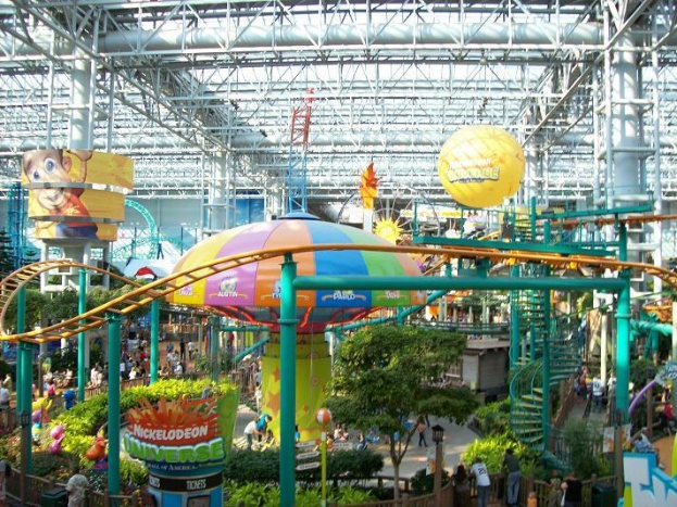 Ein großes Indoor-Wasserpark mit einer Achterbahn, umgeben von Menschen, Grünflächen und beleuchteten Deckenstrukturen.