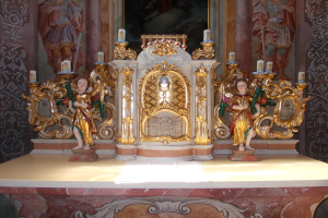 Ein Altar in einer Kirche mit Skulpturen von Menschen und Kerzen auf Ständern, mit einem Gemälde an der Wand dahinter, das eine feierliche Atmosphäre schafft.