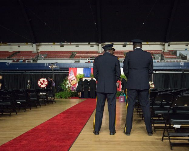 Zwei uniformierte Männer stehen auf einem roten Teppich bei einer Abschlussfeier, umgeben von einer Menge mit Blumensträußen, Stühlen, Fahnen und Pflanzen im Hintergrund.