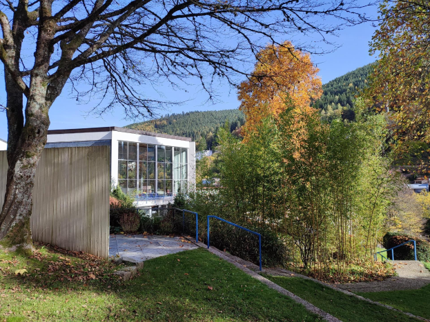 Das Universitätsgebäude Stuttgart mit einem Baum davor, umgeben von Gras, trockenen Blättern, Pflanzen und einem Geländer, mit Hügeln und einem klaren blauen Himmel im Hintergrund.