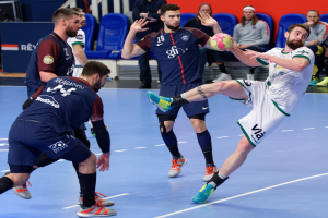 Eine Gruppe von Männern, die Handball auf einem Platz spielen, wobei einer den Ball hält, während Zuschauer von Stühlen, Geländern und einer Wand im Hintergrund zuschauen.