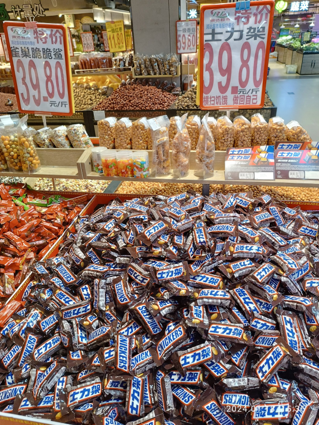 Eine Vitrine voller verschiedener Süßigkeiten, einschließlich Snickers-Riegel, in einem Geschäft mit Lebensmitteln auf Regalen, Texttafeln, Deckenleuchten und Hintergrundobjekten.