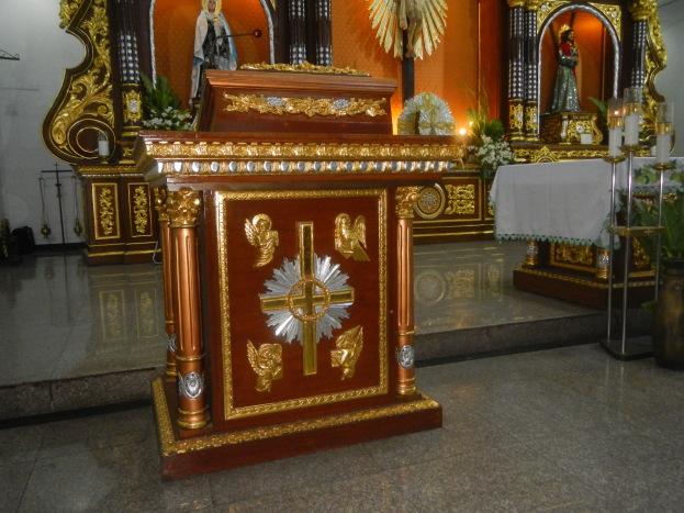 Ein Altar in einer Kirche mit einem Kreuz, Statuen, Blumensträußen, Kerzen mit Ständern, einem mit einem Tuch bedeckten Tisch und anderen Gegenständen, wobei die Wände mit Lichtern geschmückt sind.