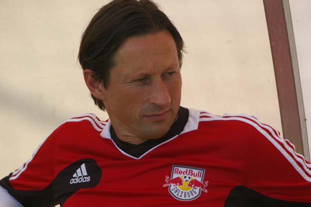 Ein Mann in einem roten und schwarzen T-Shirt sitzt auf einer Bank vor einer Wand und schaut selbstbewusst und entschlossen, wie ein professioneller Fußballspieler.