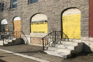 Ein Gebäude mit vernagelten Fenstern und einem Schild mit der Aufschrift "Günstiger Wohnraum", mit einer Treppe und Geländern, die nach oben führen, und einer Straße im Vordergrund.
