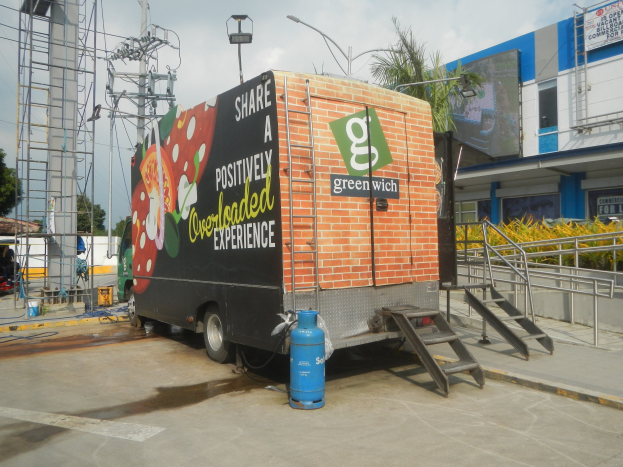 Lastwagen vor einem Gebäude mit einer Tafel, auf der "Teilen Sie eine positiv überladene Erfahrung" steht, mit Stufen, einem Zylinder auf dem Boden, Geländern, Strommasten mit Drähten, Laternenpfählen, Bäumen und einer bewölkten Himmel.