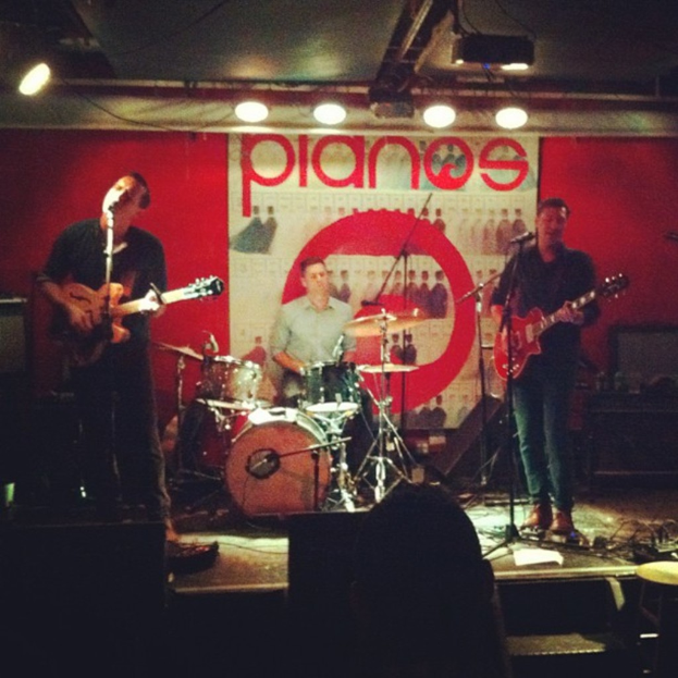 Drei Männer auf der Bühne: einer spielt Schlagzeug in der Mitte, und zwei spielen Gitarre und singen auf beiden Seiten, mit einem Banner an der Wand und Lampen an der Decke.