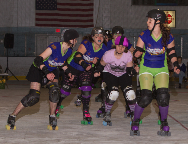 Mehrere Frauen in Eislaufschuhen und Helmen sitzen drinnen, mit einer Flagge an der Wand, einem Lautsprecher auf einem Stativ, einer Uhr und einer Tür im Hintergrund.