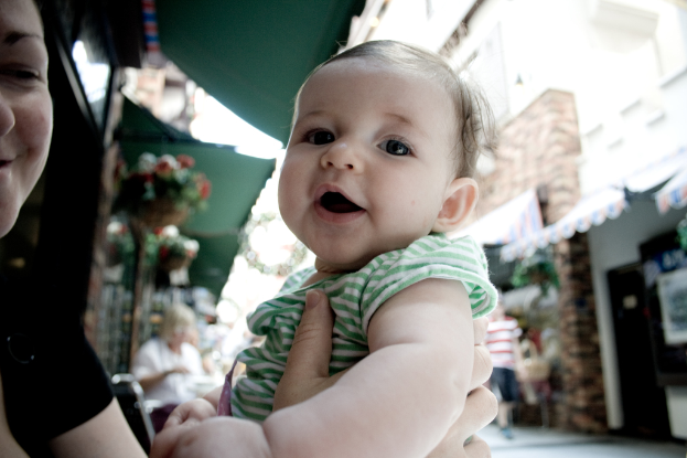 Eine Frau hält ein Kind mit einem unscharfen Hintergrund, der eine sitzende Frau, einen gehenden Mann, Hauspflanzen, Gebäude und verschiedene Gegenstände zeigt.