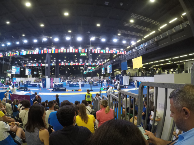 Eine große Menschenmenge auf Sitzplätzen und Stehplätzen in einer Indoor-Sportarena mit Bildschirmen, Flaggen und Deckenbeleuchtung im Hintergrund.