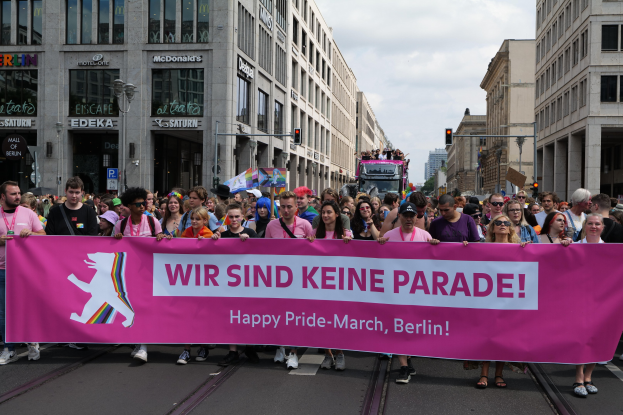 Gruppe von Menschen, die eine Straße in Berlin, Deutschland, entlanggehen und ein pinkes Banner mit der Aufschrift "Happy Pride March" halten, mit Gebäuden, Laternenmasten und Verkehrszeichen an der Straße.