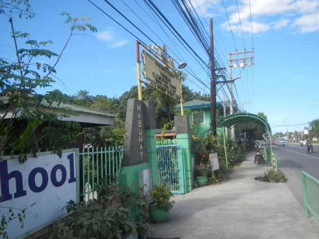 Der Eingang einer Schule mit einem Schild, auf dem "Schule" steht, auf der Seite einer Straße, mit einem Tor, einem Namenbrett, Pflanzen, Bäumen, Strommasten mit Drähten und einer bewölkten Himmel im Hintergrund.
