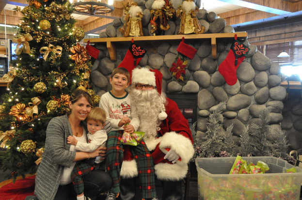 Eine Frau, Kinder und ein Mann in Santa-Klaus-Kostüm mit Brille und Handschuhen stehen neben einem geschmückten Weihnachtsbaum, einer Wand mit Felsen, Spielzeug, Dekorationen, einem Korb mit Gegenständen und Hintergrundbeleuchtung, während ein Junge ein Päckchen hält.