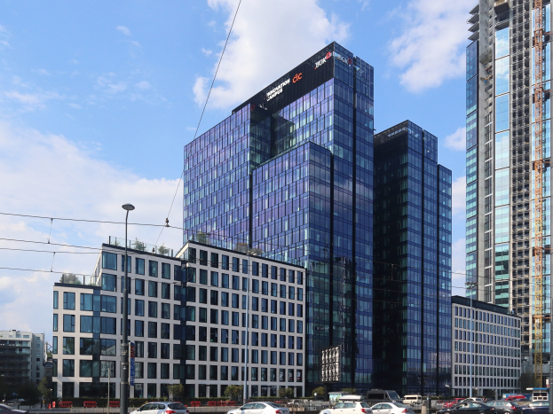 Großes modernes Gebäude mit vielen Fenstern, identifiziert als Deutsche Bank Hauptsitz in Frankfurt, Deutschland, umgeben von städtischer Infrastruktur einschließlich Straßenlaternen, Fahrzeugen, Grünflächen und einem bewölkten Himmel.