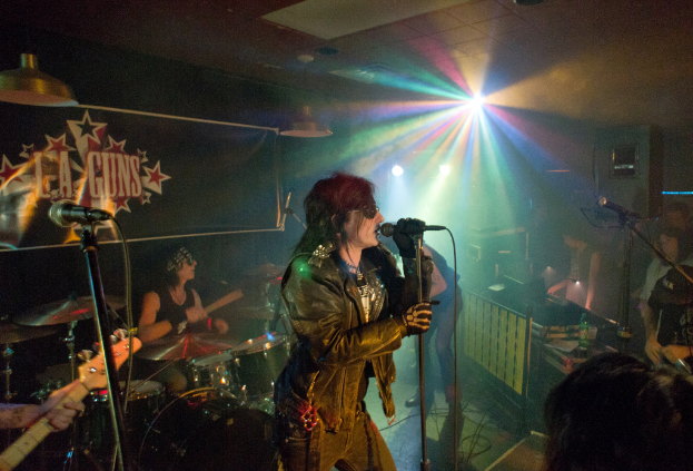 Eine Frau in einer schwarzen Jacke singt auf der BÃ¼hne in ein Mikrofon, mit einem Mann in Sonnenbrille und StirnbÃ¤nder, der hinter ihr ein Instrument spielt, und einem Plakat mit 'LA Guns' im Hintergrund.