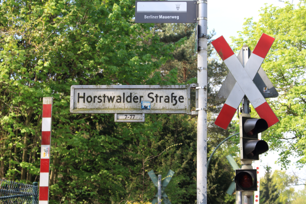 Ein Mast mit einer Ampel und einem Schild mit der Aufschrift "Horstwalder Stra├če" im Vordergrund, mit B├Ąumen im Hintergrund und einem Zaun auf der linken Seite.