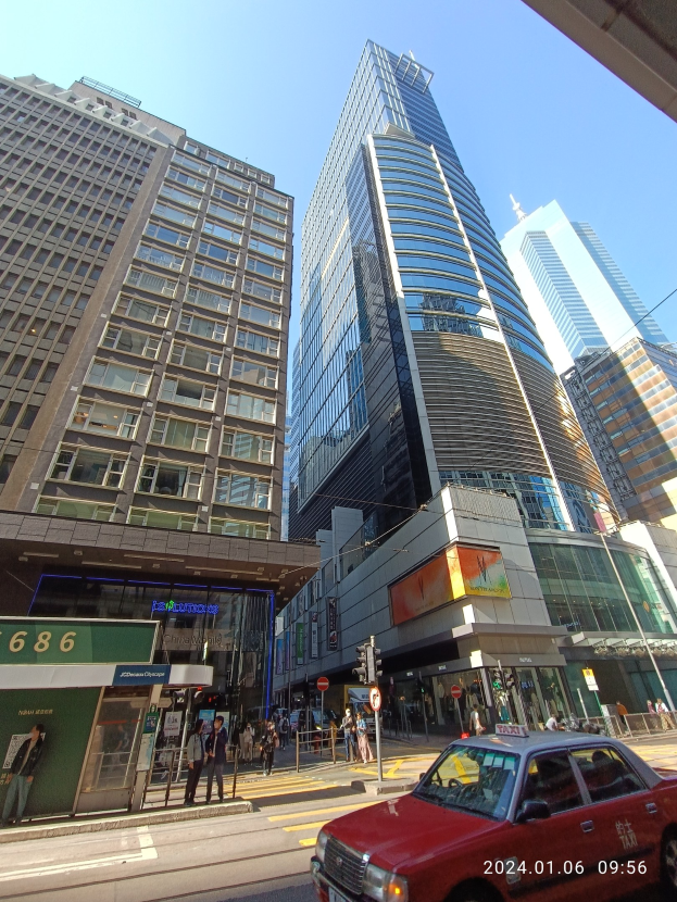 Roter Wagen auf einer Stadtstraße mit hohen Wolkenkratzern, Passanten auf dem Gehweg, Verkehrsampeln und sichtbarer Beschilderung bei klarem Himmel.