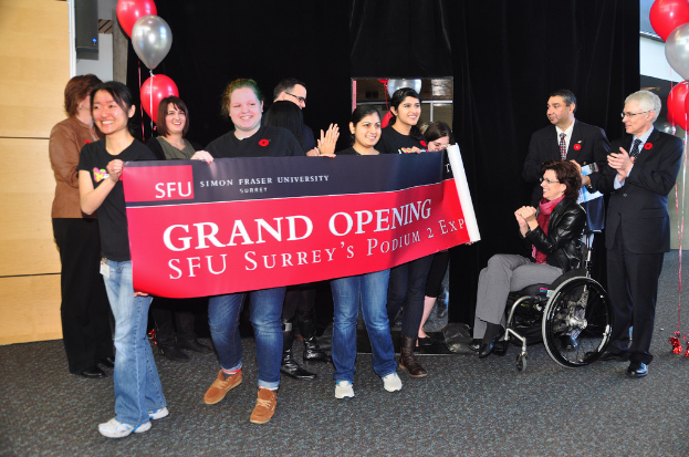 Eine Gruppe Frauen hält ein Banner hoch, zwei klatschende Männer stehen daneben und eine Frau im Rollstuhl sieht begeistert aus, vor einem schwarzen Vorhang mit Ballons.