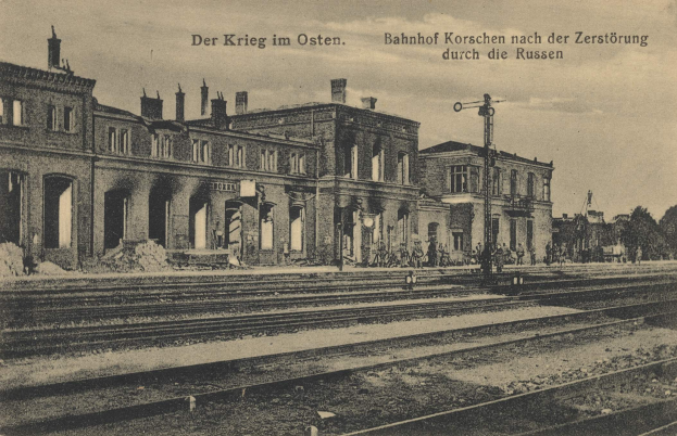 Schwarß-weißes Foto der Bahnstation Korschen in Ostern, Deutschland, mit Gleisen, Stationsgebäuden, Strommasten, Menschen, Bäumen und Himmel.