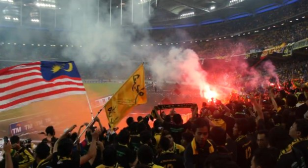 Große Menge in einem Stadion mit Fackeln, Fahnen, Rauch und Schildern mit der Aufschrift "Malaysia vs Malaysia Live-Streaming" bei einem Ereignis.