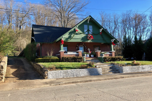 Ein gr├╝nes Haus mit einem Kranz an der Vorderseite, umgeben von Pflanzen, Gras, Treppen, einer Wand, B├Ąumen und Dr├Ąhten, unter einem bew├Âlktem Himmel, im Herzen der Stadt und zur Miete verf├╝gbar.