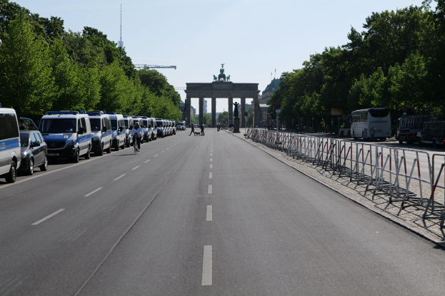 Lange Reihe von Polizeiautos, die auf der Straße vor dem Brandenburger Tor in Berlin, Deutschland, geparkt sind, mit Menschen auf Fahrrädern und in der Nähe stehenden, Barrieren, Bäumen und einem Bogen mit Statuen im Hintergrund.