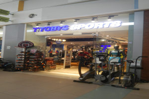 Ladenfront von "Toby's Sports" mit Schaufensterpuppen, Fitnessgeräten und einer Tafel mit lesbarem Text im Inneren.