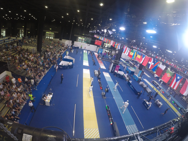 Große Halle mit Zuschauern bei einer Gymnastikveranstaltung, Sitzplätze auf der linken und rechten Seite, Stehende auf dem Boden, Deckenbeleuchtung und Flaggen im Hintergrund.