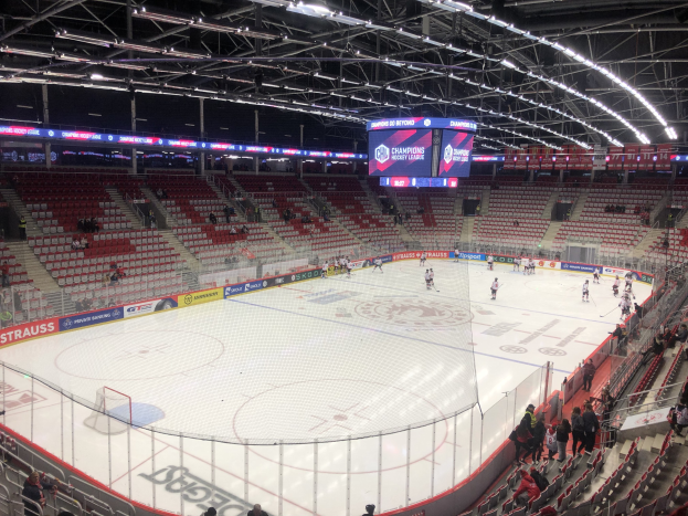 Eishockeyspiel in einer großen Arena mit Zuschauern, eine von einem Zaun umgebene Eisfläche, ein Display, das "St. Louis Blues gegen Detroit Red Wings" anzeigt, Metallrahmen, Deckenleuchten und ein Dach.